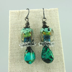 Peut inclure: Paire de boucles d'oreilles artisanales. Chaque boucle d'oreille présente une perle rectangulaire transparente avec des accents verts et bleus, un accent argenté et une pierre turquoise en forme de goutte. Les boucles d'oreilles ont des crochets en métal foncé et un fil enroulé.