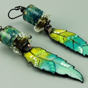 Peut inclure: Paire de boucles d'oreilles artisanales. Chaque boucle d'oreille présente une perle cylindrique en verre avec des tourbillons bleus et verts, des cristaux transparents et du fil de fer. Les boucles d'oreilles ont un pendentif en forme de plume avec de l'émail jaune et turquoise, et de petits points blancs.