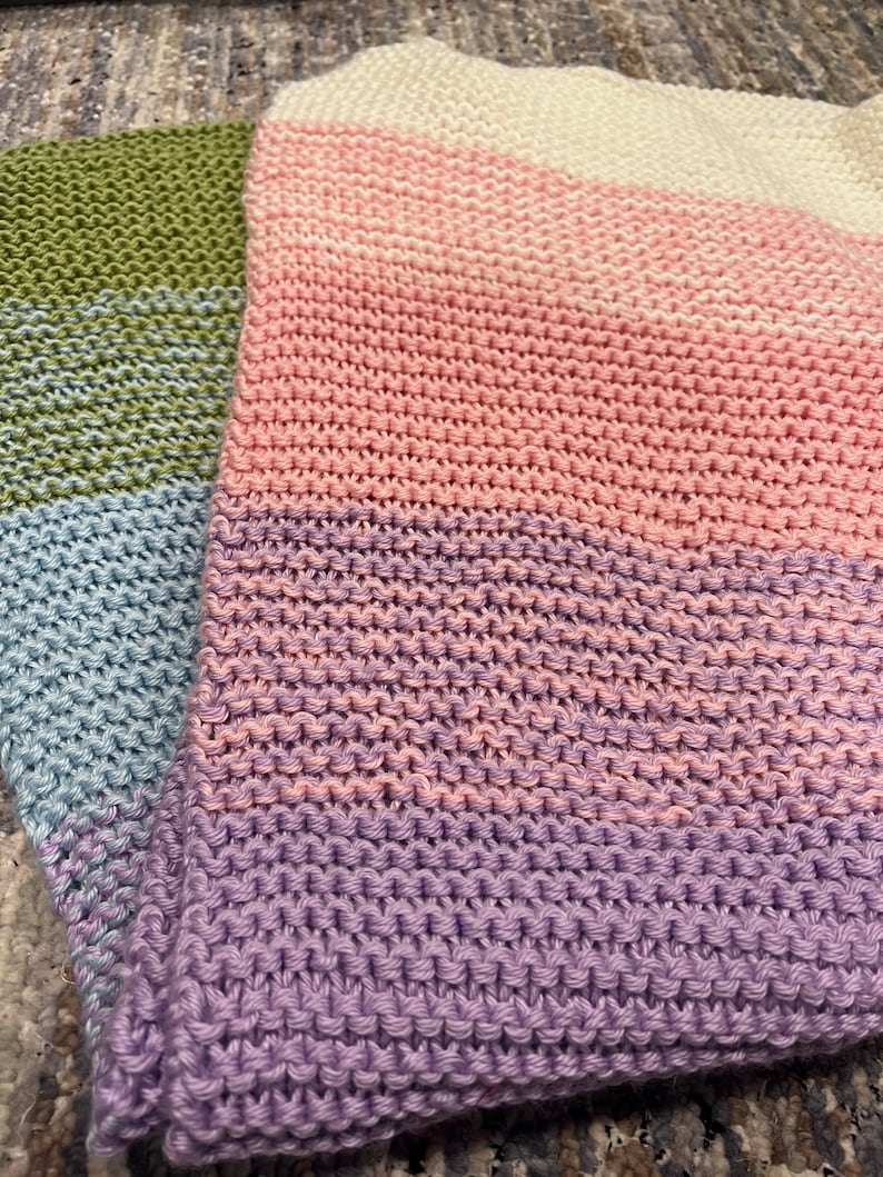 K&ouml;nnte beinhalten: Eine mehrfarbige gestrickte Decke mit Streifen in Gr&uuml;n, Blau, Rosa und Lila.