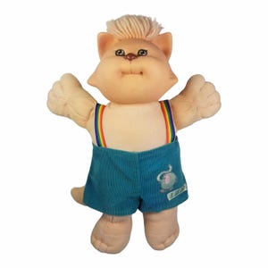 Peut inclure: Une poupée en peluche en forme de chat avec des cheveux blonds, portant une salopette bleue avec des bretelles arc-en-ciel. La poupée a un visage souriant avec des yeux bruns et un nez rose. La salopette a un petit motif brodé de chat sur le devant.
