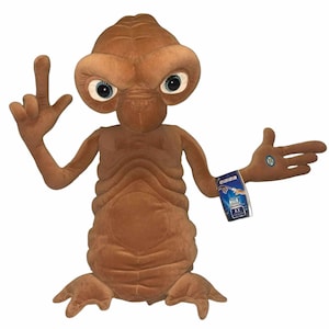 Puede incluir: Un juguete de peluche marrón del personaje extraterrestre E.T. de la película E.T., el extraterrestre. El juguete está de pie con los brazos extendidos, una mano haciendo un signo de paz y la otra apuntando hacia un lado. El juguete tiene ojos grandes y una expresión amigable. El juguete lleva una etiqueta azul que dice "E.T."