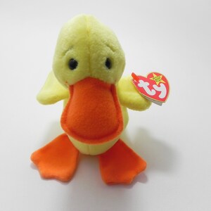 May include: A yellow plush duck toy with an orange beak and feet. The duck has black eyes and small wings. A red heart-shaped tag with the text "TY" is attached to the duck.