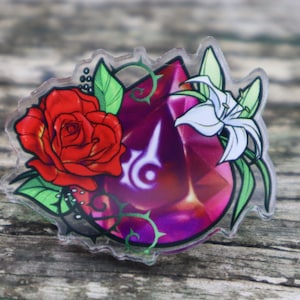 Könnte beinhalten: Eine durchsichtige Acryl-Anstecknadel mit einem violetten Edelstein-Design, einer roten Rose und einer weißen Lilie. Der Edelstein hat ein stilisiertes Symbol in der Mitte.