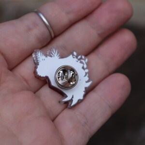May include: White enamel pin featuring a detailed design of a stylised floral arrangement. The pin has a silver metal backing with a safety clasp.