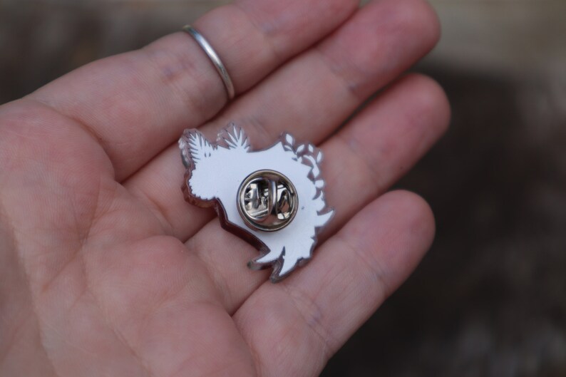 May include: White enamel pin featuring a detailed design of a stylised floral arrangement. The pin has a silver metal backing with a safety clasp.