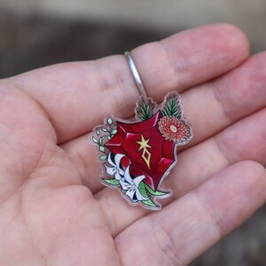 May include: A red gemstone charm with a gold star and diamond symbol, surrounded by white flowers and green leaves. The charm is attached to a clear acrylic keyring.