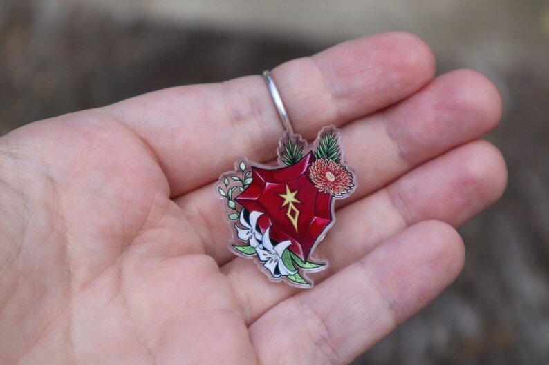 May include: A red gemstone charm with a gold star and diamond symbol, surrounded by white flowers and green leaves. The charm is attached to a clear acrylic keyring.