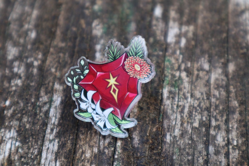 May include: A red, crystal-shaped pin with a white star and a geometric design inside. The pin is surrounded by white flowers and green leaves.