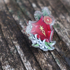 May include: A red, star-shaped, clear acrylic pin with a white flower design. The pin is on a wooden surface.
