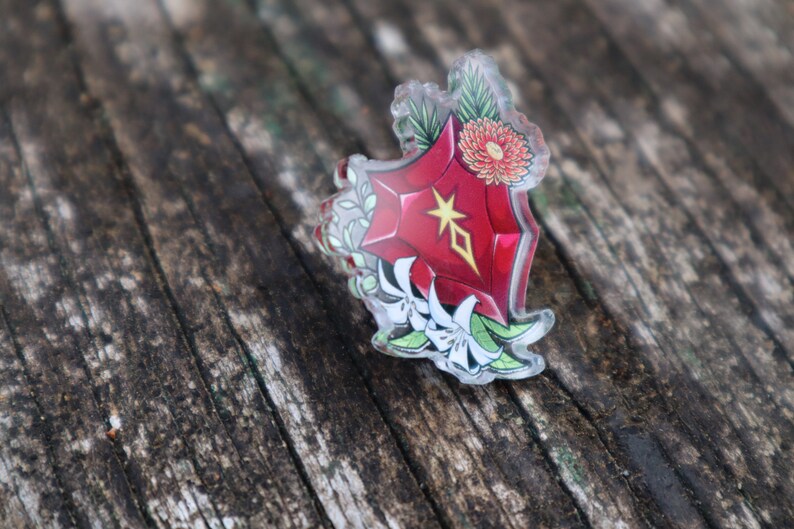 May include: A red, star-shaped, clear acrylic pin with a white flower design. The pin is on a wooden surface.
