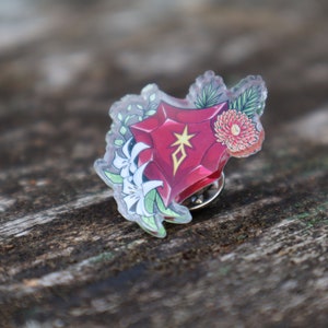 May include: A red, clear acrylic pin with a floral design. The pin features a red gem with a gold star and a white flower.