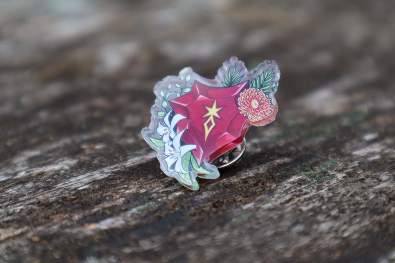 May include: A red, clear acrylic pin with a floral design. The pin features a red gem with a gold star and a white flower.
