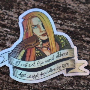 Puede incluir: Una pegatina holográfica con una mujer de pelo largo y rojo y un atuendo verde y dorado. La pegatina tiene una pancarta con el texto "I will set this world ablaze And we shall dance before the pyre."