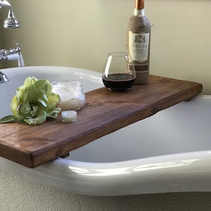 May include: A wooden bath tray with a glass of red wine, a candle, and a jar of bath salts. The tray is placed on the edge of a white bathtub.