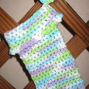 May include: A crocheted dishcloth in shades of green, purple, blue, and white. The dishcloth is hanging on a wooden lattice.