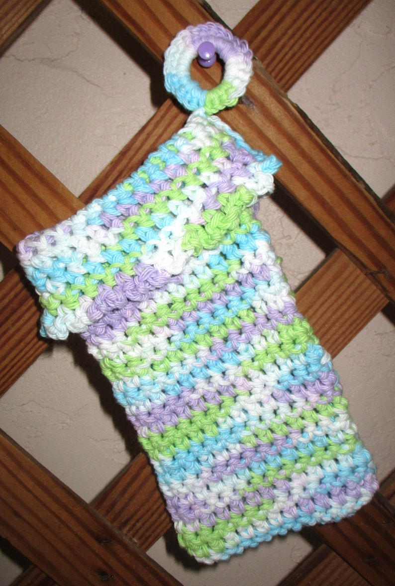 May include: A crocheted dishcloth in shades of green, purple, blue, and white. The dishcloth is hanging on a wooden lattice.