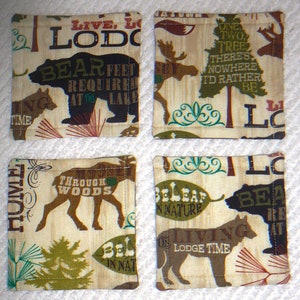 Puede incluir: Cuatro posavasos de tela beige con costuras marrones y varios dise&ntilde;os con tem&aacute;tica de la naturaleza. Los posavasos presentan texto como "Live, Lodge", "Bear Feet Required at the Lake", "One Two Tree There's Nowhere I'd Rather Be", "Through Woods", "Believe in Nature", y "Living on Lodge Time".