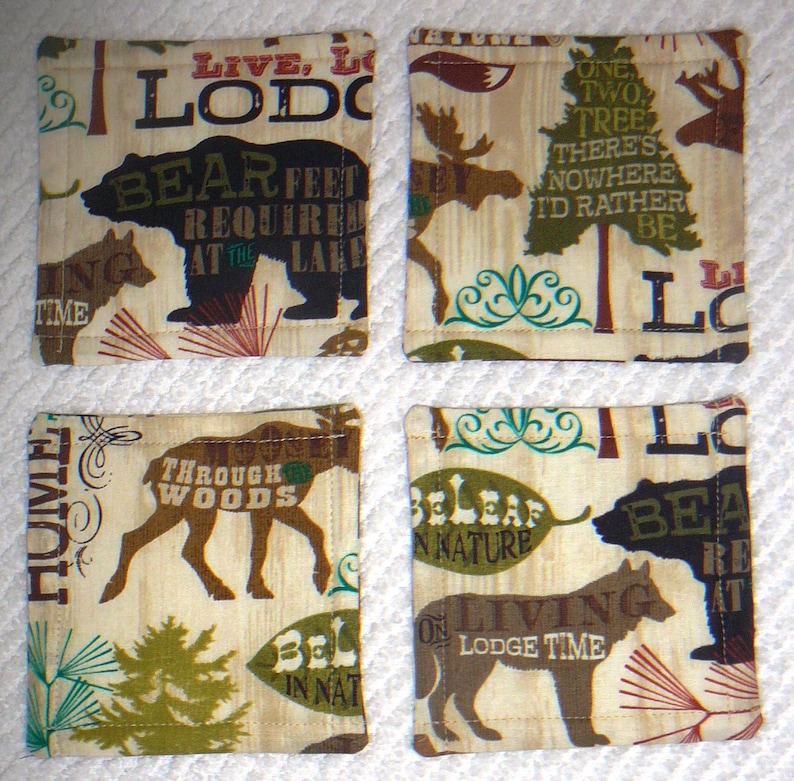 Puede incluir: Cuatro posavasos de tela beige con costuras marrones y varios dise&ntilde;os con tem&aacute;tica de la naturaleza. Los posavasos presentan texto como "Live, Lodge", "Bear Feet Required at the Lake", "One Two Tree There's Nowhere I'd Rather Be", "Through Woods", "Believe in Nature", y "Living on Lodge Time".