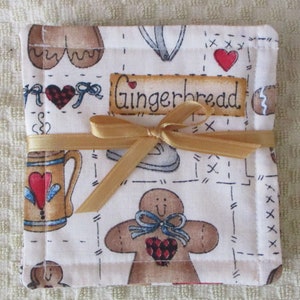 Puede incluir: Un posavasos de tela blanca con tem&aacute;tica de pan de jengibre. El posavasos presenta un hombre de jengibre, una taza y un coraz&oacute;n con la palabra "Gingerbread" impresa en &eacute;l. El posavasos est&aacute; atado con una cinta dorada.