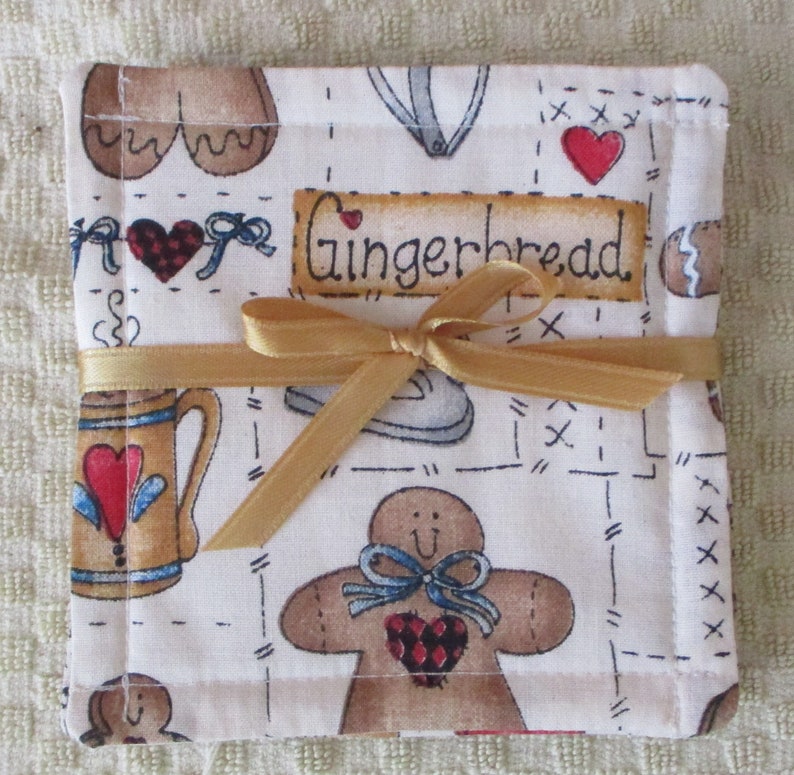 Puede incluir: Un posavasos de tela blanca con tem&aacute;tica de pan de jengibre. El posavasos presenta un hombre de jengibre, una taza y un coraz&oacute;n con la palabra "Gingerbread" impresa en &eacute;l. El posavasos est&aacute; atado con una cinta dorada.
