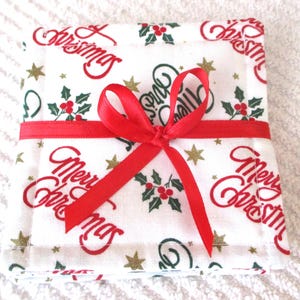 Puede incluir: Un juego de cuatro posavasos de tela blanca con una cinta roja atada alrededor. Los posavasos tienen un patr&oacute;n repetitivo de "Feliz Navidad" en letras rojas, ramas de acebo verdes y estrellas doradas.