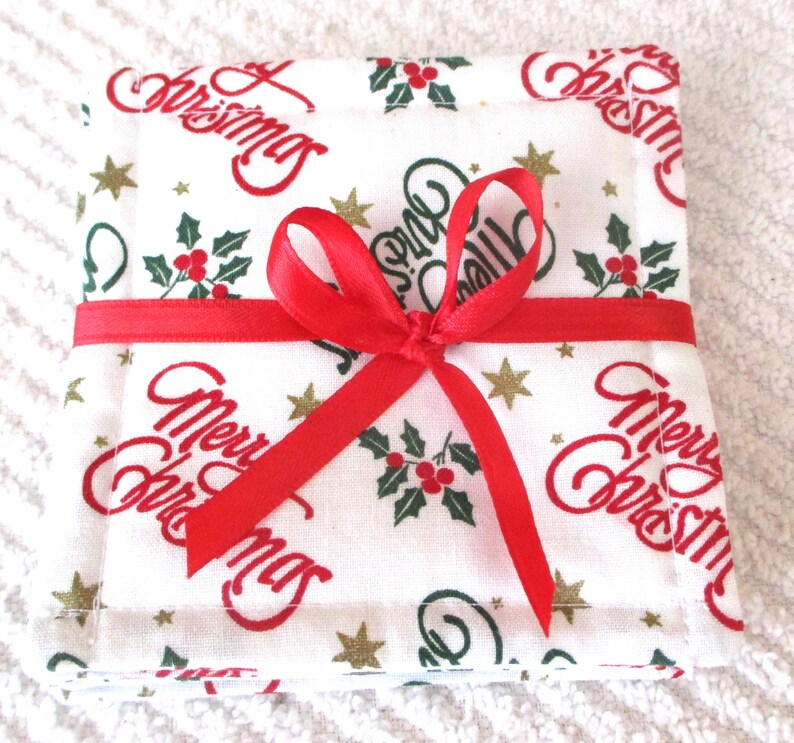 Puede incluir: Un juego de cuatro posavasos de tela blanca con una cinta roja atada alrededor. Los posavasos tienen un patr&oacute;n repetitivo de "Feliz Navidad" en letras rojas, ramas de acebo verdes y estrellas doradas.