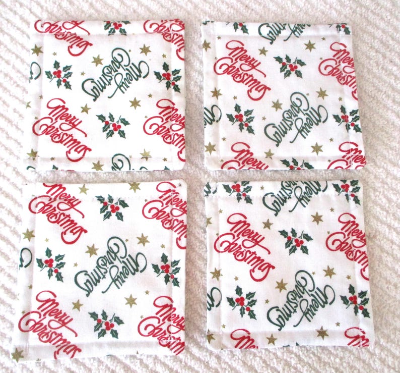 Puede incluir: Conjunto de cuatro posavasos de tela blanca con un estampado navide&ntilde;o rojo y verde. El estampado presenta las palabras "Feliz Navidad" y "Felices Fiestas" con acebo y estrellas doradas.