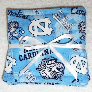Puede incluir: Un juego de cuatro posavasos de tela azul y blanco con un logotipo de UNC Tar Heels y las palabras "Tar Heels" y "North Carolina". Los posavasos están atados con una cinta blanca.