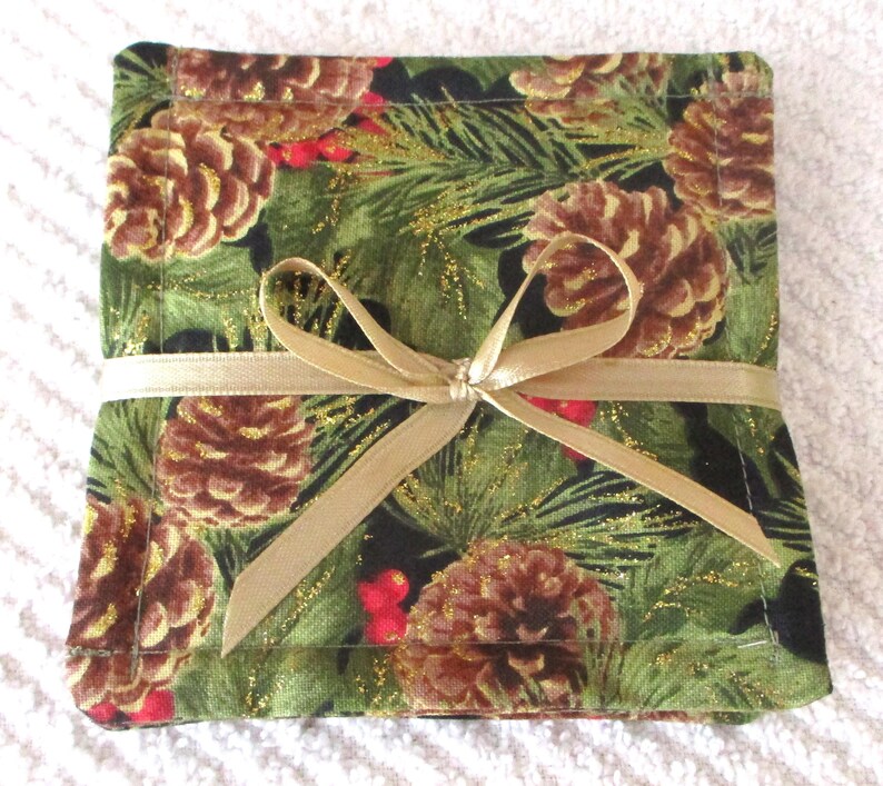 Puede incluir: Conjunto de cuatro posavasos de tela verde y negro con un patr&oacute;n de pi&ntilde;as y bayas de acebo. Los posavasos est&aacute;n atados con una cinta dorada.