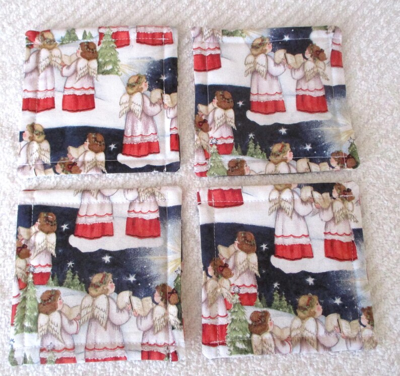 Puede incluir: Conjunto de cuatro posavasos cuadrados de tela con un dise&ntilde;o de &aacute;ngel navide&ntilde;o. Los posavasos son blancos con un fondo azul oscuro y presentan &aacute;ngeles cantando en un paisaje nevado. Los &aacute;ngeles llevan t&uacute;nicas blancas con detalles rojos y tienen halos dorados.