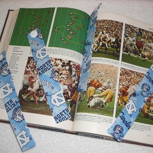 Peut inclure: Quatre signets en tissu bleu et blanc avec le texte "North Carolina" et "UNC" dessus. Les signets sont plac&eacute;s sur un livre ouvert avec des photos de joueurs de football.