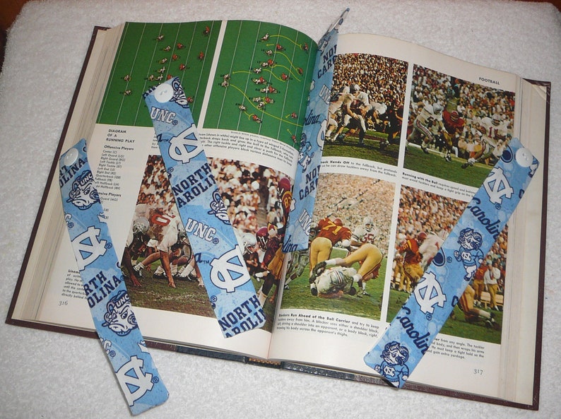Peut inclure: Quatre signets en tissu bleu et blanc avec le texte "North Carolina" et "UNC" dessus. Les signets sont plac&eacute;s sur un livre ouvert avec des photos de joueurs de football.
