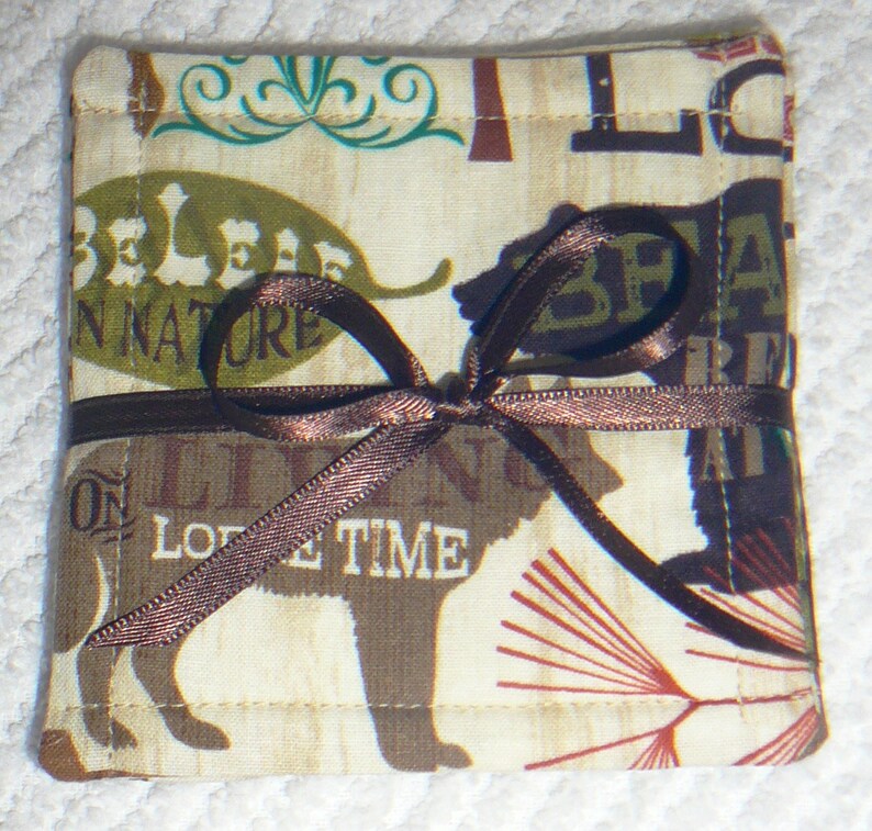 Puede incluir: Un posavasos de tela marr&oacute;n y beige con una cinta marr&oacute;n atada alrededor. El posavasos tiene un tema de naturaleza con las palabras "Believe in Nature" y "On Love Time" impresas en &eacute;l. El posavasos tambi&eacute;n presenta una silueta de oso marr&oacute;n.