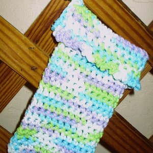 May include: A crocheted dishcloth in shades of green, blue, purple, and white. The dishcloth is hanging on a wooden lattice.