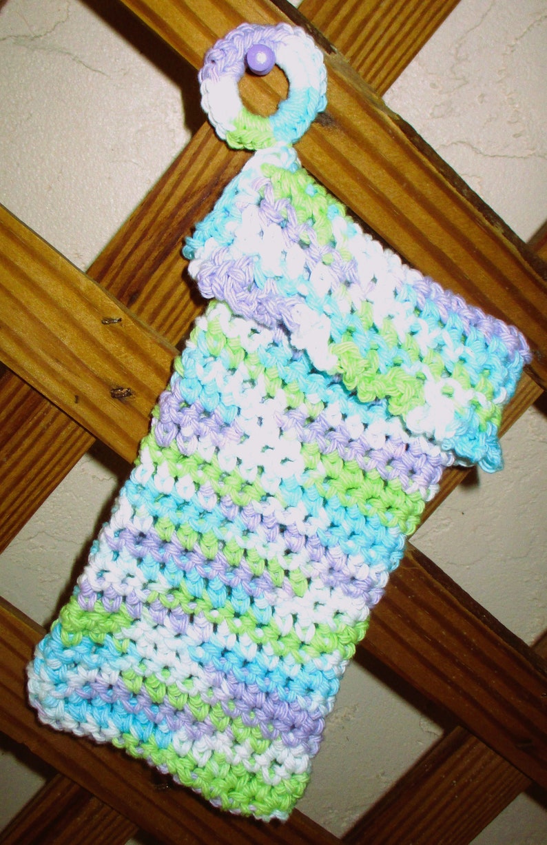 May include: A crocheted dishcloth in shades of green, blue, purple, and white. The dishcloth is hanging on a wooden lattice.