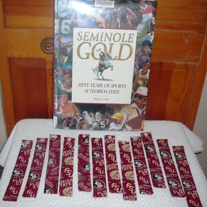 May include: A book titled "Seminole Gold: Fifty Years of Sports at Florida State" by Philip L. Ben, with a maroon and gold cover featuring images of athletes and a Seminole Indian riding a horse.  The book is surrounded by 14 maroon and gold fabric bookmarks with the Florida State University logo.