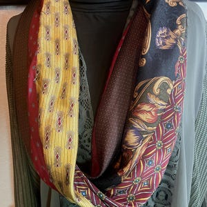 May include: A multi-patterned infinity scarf. The scarf features sections of yellow with brown diamond shapes, burgundy with small dots, and black with floral designs. The scarf is made of a silky material and is draped around a black turtleneck.