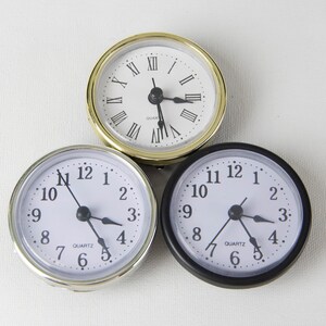 May include: Three round analog clocks with white faces and black numbers. The clock on the left has a silver rim, the clock in the middle has a gold rim, and the clock on the right has a black rim.