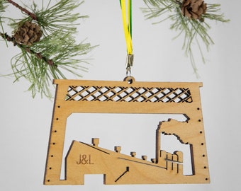 May include: A wooden ornament shaped like a factory with a smokestack and the text "J&L" on the side. The ornament is hanging from a green and yellow ribbon.