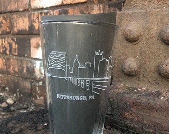 May include: A clear glass pint glass with an etched design of the Pittsburgh skyline and the words "Pittsburgh, PA".