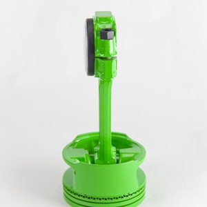 Op de afbeelding: Een groen geverfde zuiger met een zwarte ring en een groene drijfstang. De zuiger staat op een wit oppervlak.