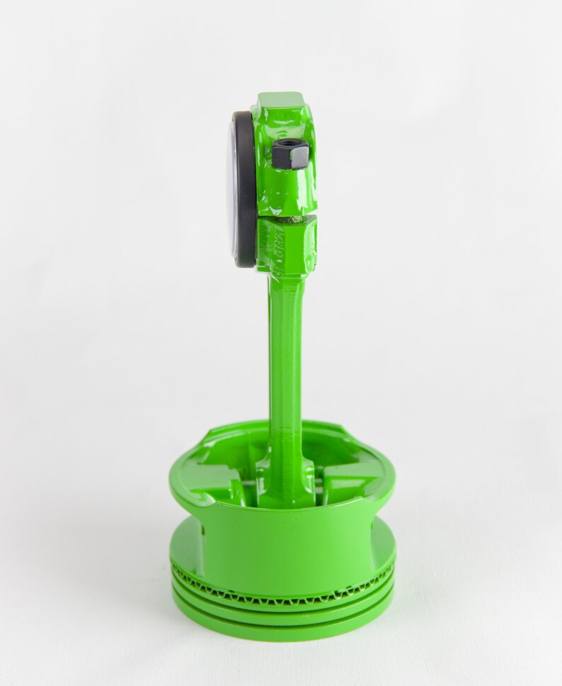 Op de afbeelding: Een groen geverfde zuiger met een zwarte ring en een groene drijfstang. De zuiger staat op een wit oppervlak.