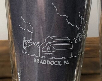 May include: Clear glass tumbler with a detailed white outline of a factory and the text "BRADDOCK, PA". The glass contains a dark gray folded paper. The glass is sitting on a wooden surface.