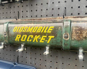 May include: A green metal valve cover with the words "OLDSMOBILE ROCKET" in yellow lettering. The valve cover is mounted on a pegboard wall with several spark plugs attached to it.