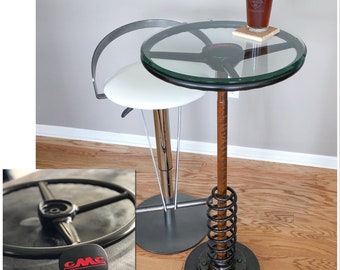 May include: A black and silver bar stool with a white seat and a glass top table with a spring base. The table has a glass top with a steering wheel design. A glass of iced tea sits on a coaster on the table.
