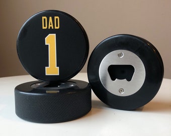 May include: Two black hockey pucks with a bottle opener. One puck has the word "DAD" and the number "1" in yellow.