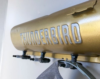 May include: A gold-colored wall-mounted coat rack with the word "THUNDERBIRD" in silver lettering. The rack features three spark plug hooks and is holding a gray and white patterned hat.