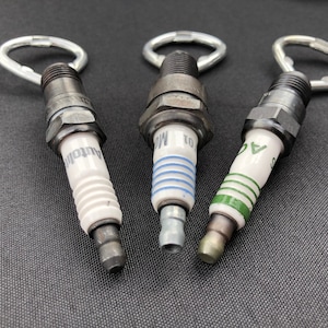May include: Three spark plugs with silver keychains attached. The spark plugs are white with black and green accents. The spark plugs are labeled "Autolite", "NGK", and "AC".