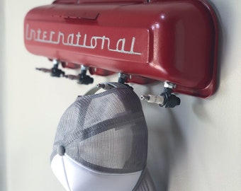 May include: A red metal wall-mounted hat rack with the word "International" in silver script. A gray and white trucker hat hangs from the rack. The rack has multiple hooks for hanging hats.