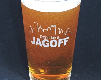 May include: A tall glass filled with a light amber colored beverage. The glass has a white cityscape design and the text "Don't be a JAGOFF" printed on it.